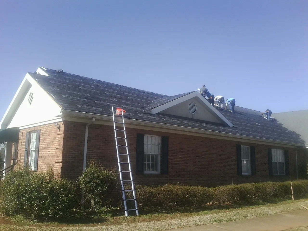 Crew tearing shingles off a brick ranch roof with a ladder set — Metal Roof Repair work by our crew in North